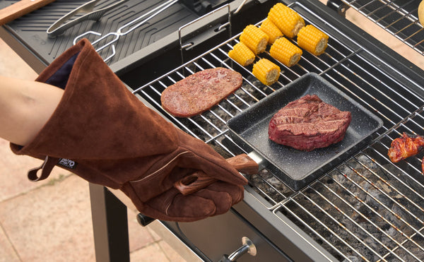 Leren BBQ handschoen met lange mouw houdt een grillpan met steak boven de gloeiende houtskool, met mais en een burger op het rooster ernaast